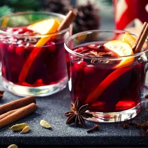 Glogg Swedish mulled wine in clear glass mugs with cranberries, orange slices, and cinnamon sticks. Cardamon pods and star anise on the table.