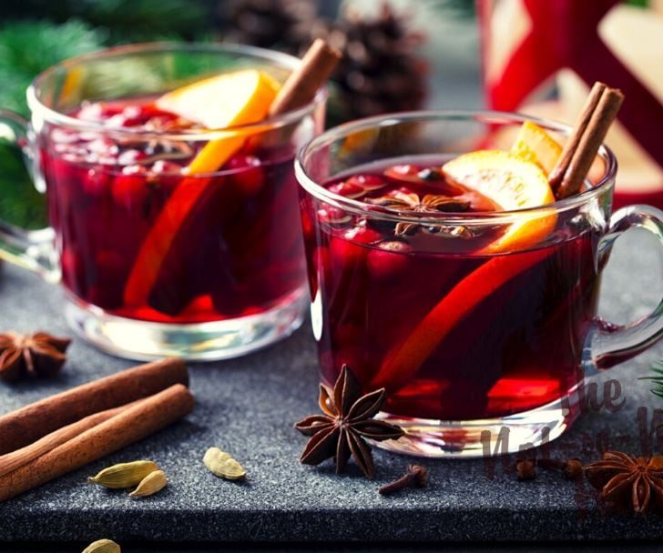 Glogg Swedish mulled wine in clear glass mugs with cranberries, orange slices, and cinnamon sticks. Cardamon pods and star anise on the table.