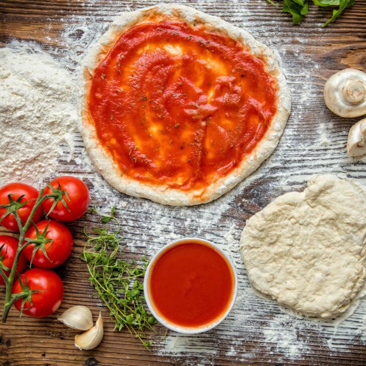 pizza dough rolled out and covered in sauce on a wood surface dusted with flour - surrounded by pizza ingredients